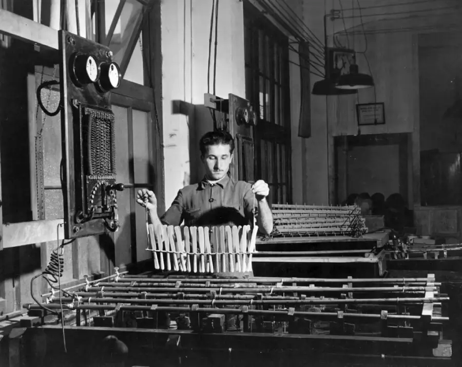A person operates machinery in a factory, monitoring gauges and handling metal rods or wires. The setting appears industrial, with visible equipment, cables, and dim lighting.