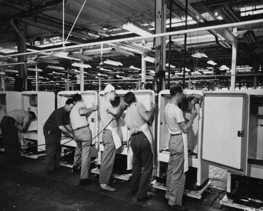 Factory workers assemble refrigerators on a production line; each person is focused on a separate unit in the busy, industrial factory setting, with overhead lights illuminating the workspace.