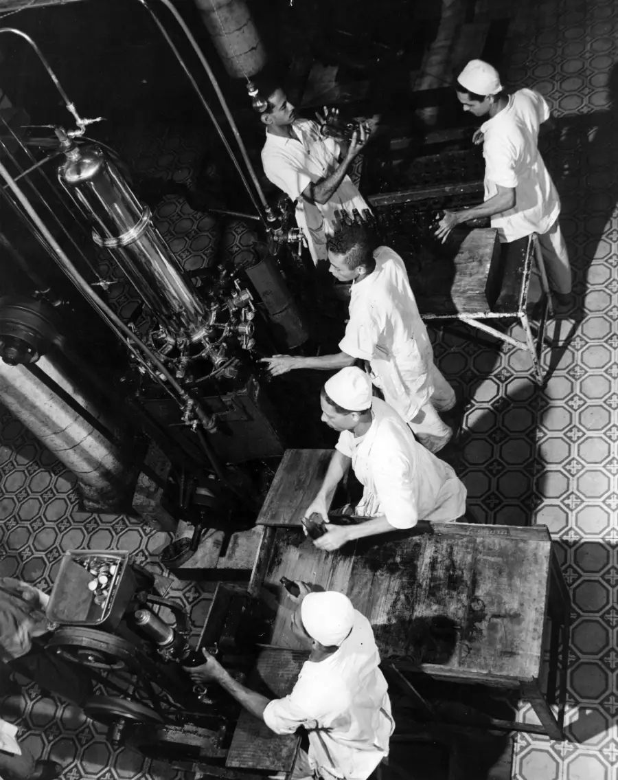 Five workers in white uniforms and caps operate machinery in a factory with tiled floors, viewed from above. Some workers handle equipment, while others attend to worktables. The scene suggests an industrial or food processing setting.