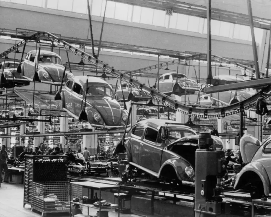 Black and white photo of a car factory assembly line with multiple Volkswagen Beetles being moved overhead and assembled below by workers.