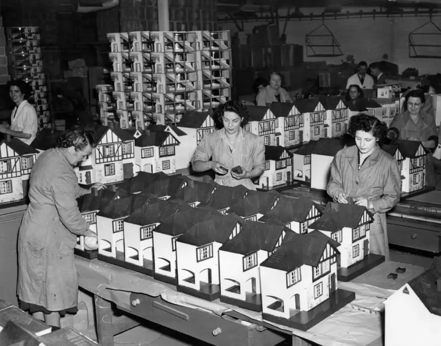 Women in a factory assemble rows of model houses on worktables, with more models stacked in the background. The scene shows teamwork in a busy workshop setting.