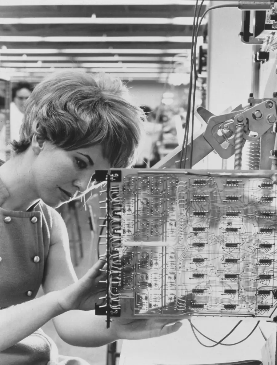 A woman with short hair works on wiring a large circuit board in a lab or factory setting, focusing intently on the electronic components.