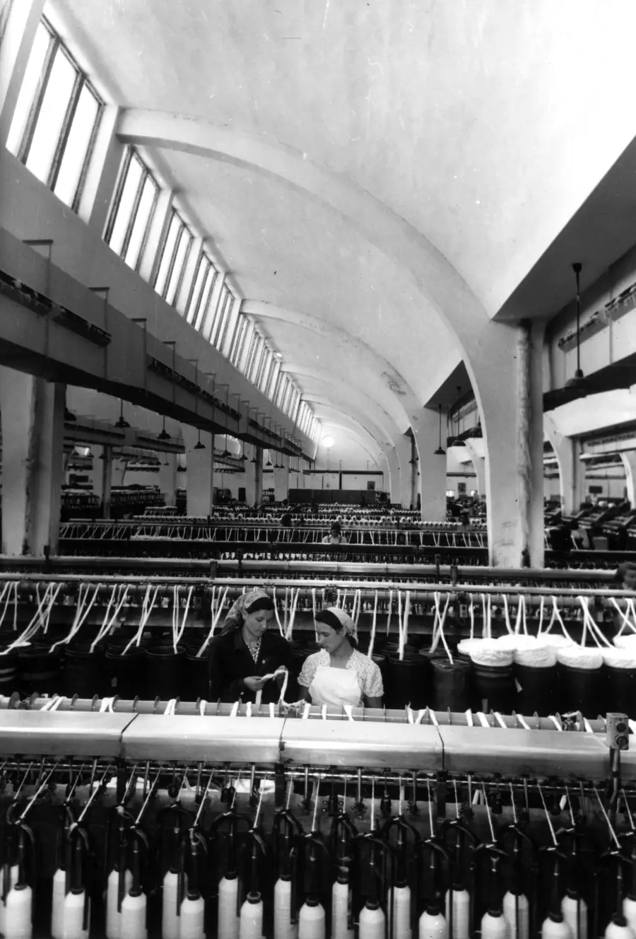 Two workers stand among rows of spinning machines inside a large, bright textile factory with arched ceilings and high windows.
