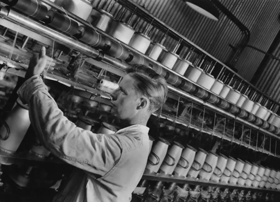 A man operates a large, industrial textile machine, adjusting spools of thread in a factory setting with rows of equipment and yarn visible. Black and white photo.