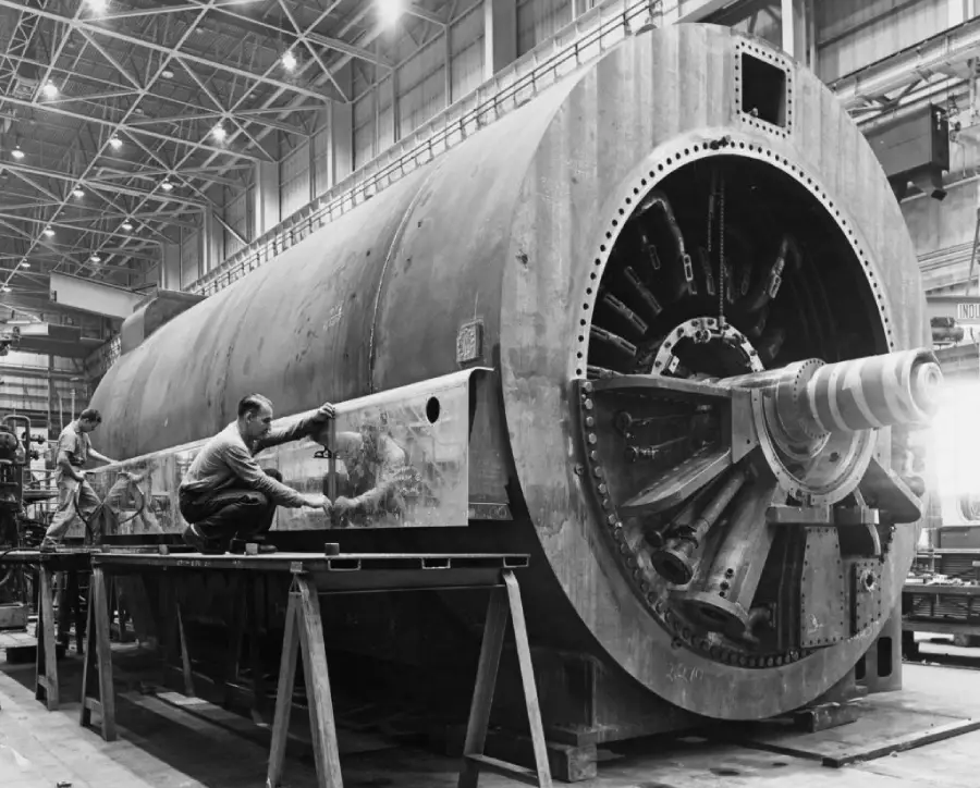 Black and white photo of two workers inspecting or assembling a large industrial machine or generator inside a spacious factory, surrounded by metal beams and industrial equipment.