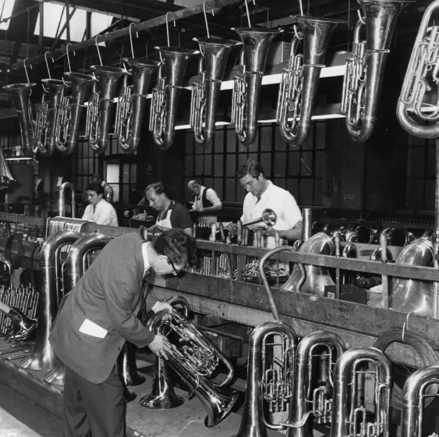 Black and white photo of workers inspecting and polishing tubas in a brass instrument factory, with many tubas hanging above and lined up below on workbenches.
