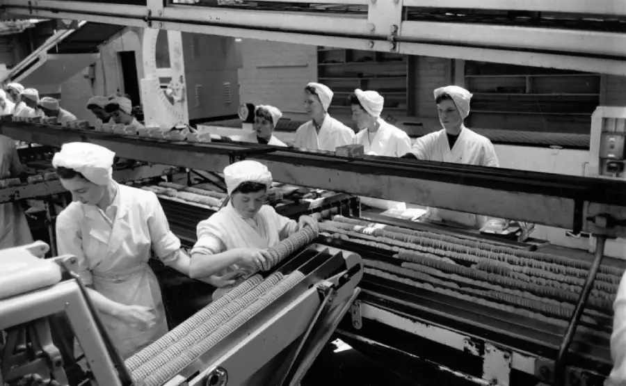 Workers in white uniforms and hair coverings assemble cookies or biscuits on a conveyor belt in a factory setting. Several women focus on sorting or inspecting items as they move along the production line.