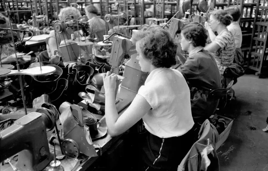 Women sit in rows at sewing machines in a busy factory, working on assembling shoes. Shelves with shoe parts surround them. The scene appears industrious and is in black and white, suggesting a historical context.