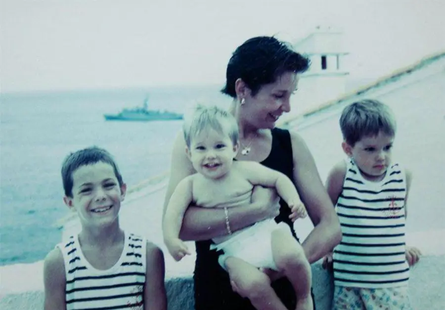 A woman holding a baby, standing with two young boys in striped shirts near a seaside wall. A ship is visible in the distance on the water. All are smiling except the boy on the right.
