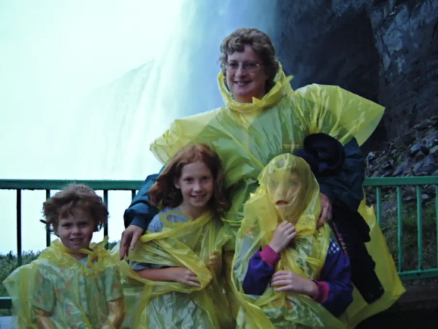 A woman and three children in yellow rain ponchos stand smiling by a railing near a waterfall, with mist and rocks in the background.