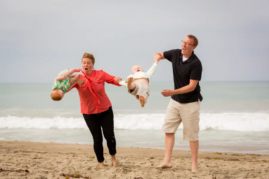 A surprised woman and laughing man swing two small children by their arms on a sandy beach with waves in the background.