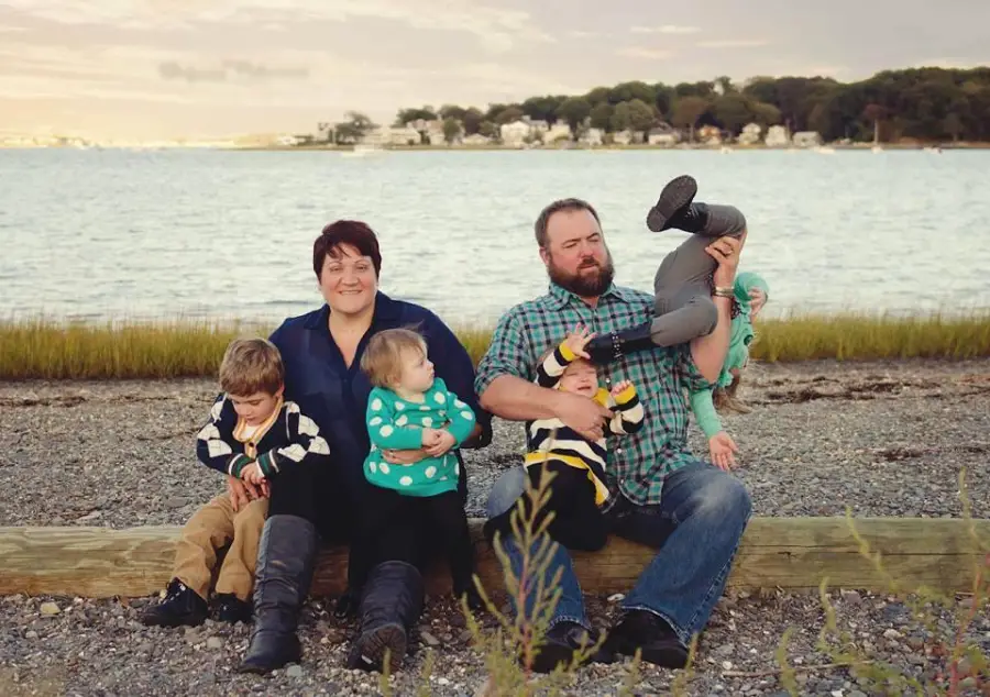 A family of five sits by a lakeshore. The mother and two kids sit calmly while the father struggles to hold two playful children, one upside down. Houses and trees are visible in the background.