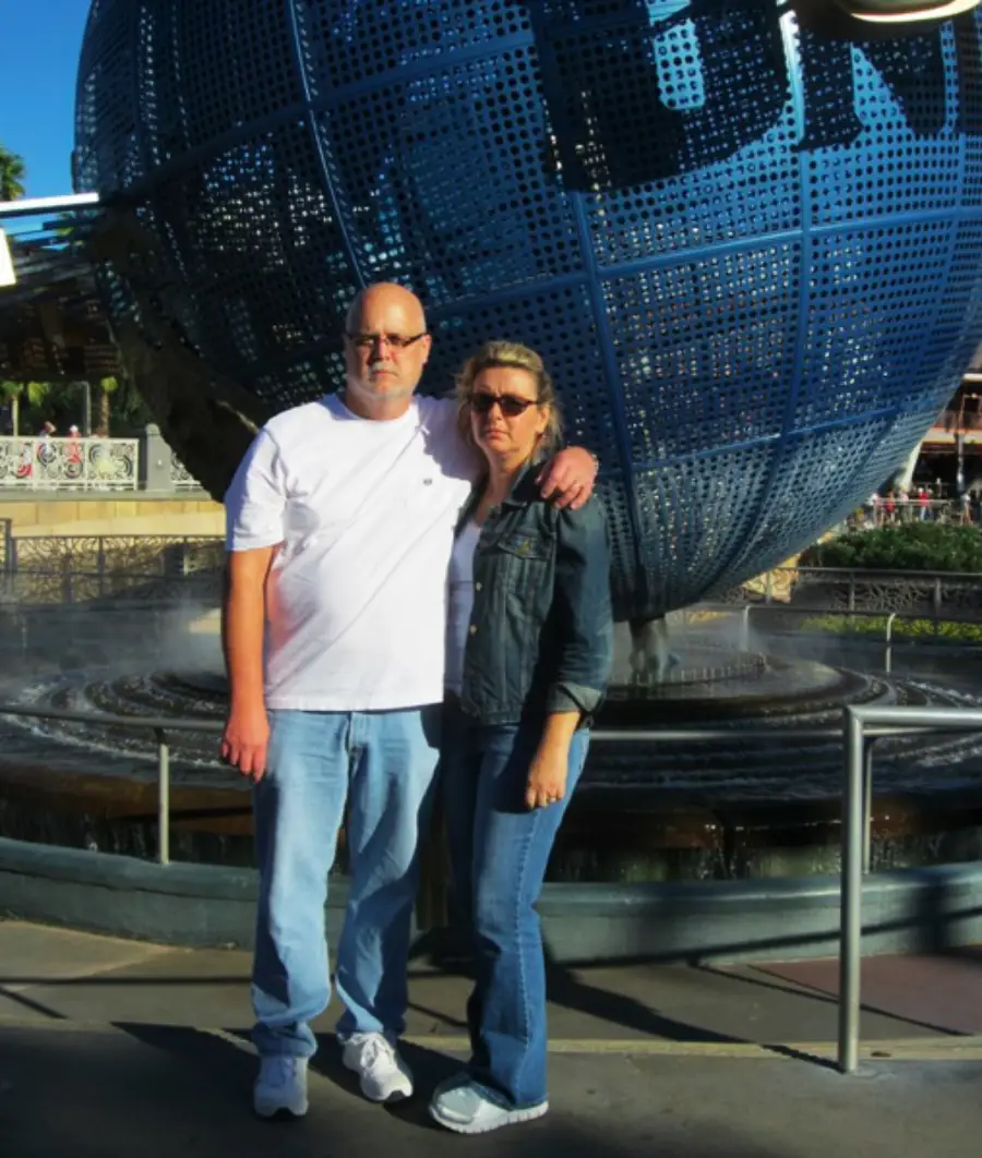 A man and woman stand together in front of a large globe fountain at Universal Studios, both wearing sunglasses and casual clothes.