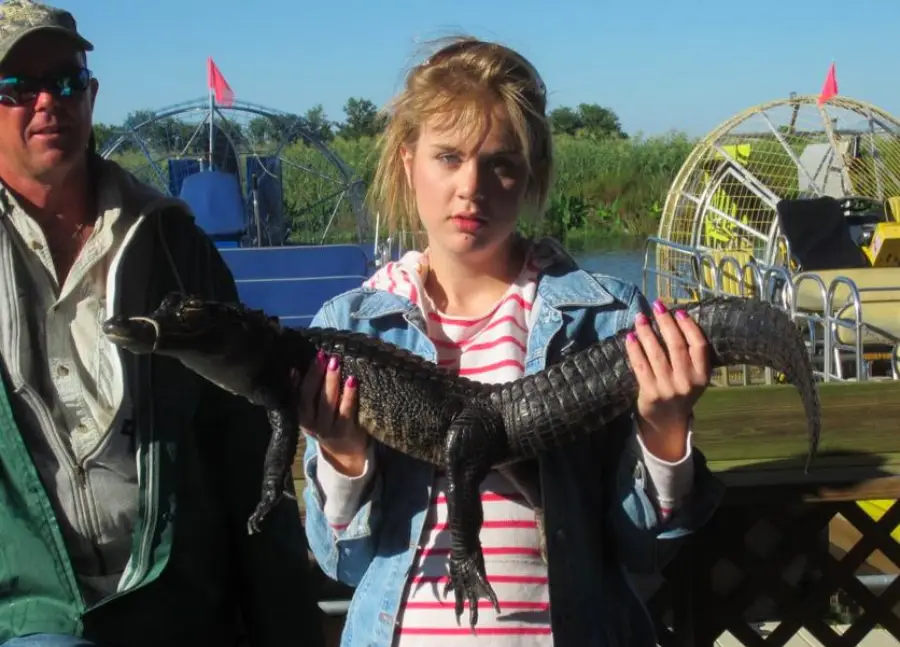 A young woman in a striped shirt and denim jacket holds a small alligator. A man in sunglasses stands nearby. Airboats and greenery are in the background.