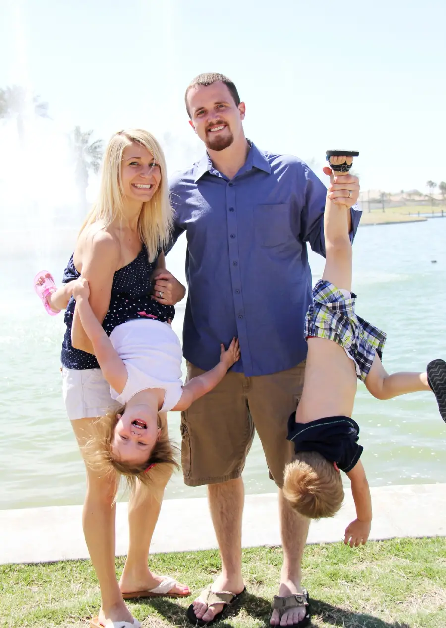 A smiling couple stands by a lake, each holding a giggling child upside down by the legs. The family appears happy, enjoying a sunny day outdoors together.