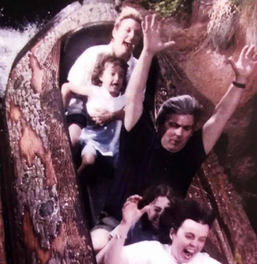 Four people ride a log flume on a water ride. The front two raise their arms, screaming with excitement, while the two behind them cling together, looking shocked and scared. Water splashes around them.