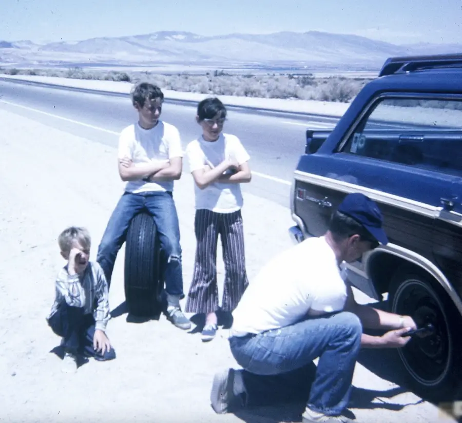 A man changes a tire on a blue station wagon while three children watch by the roadside on a sunny day in a desert-like area.