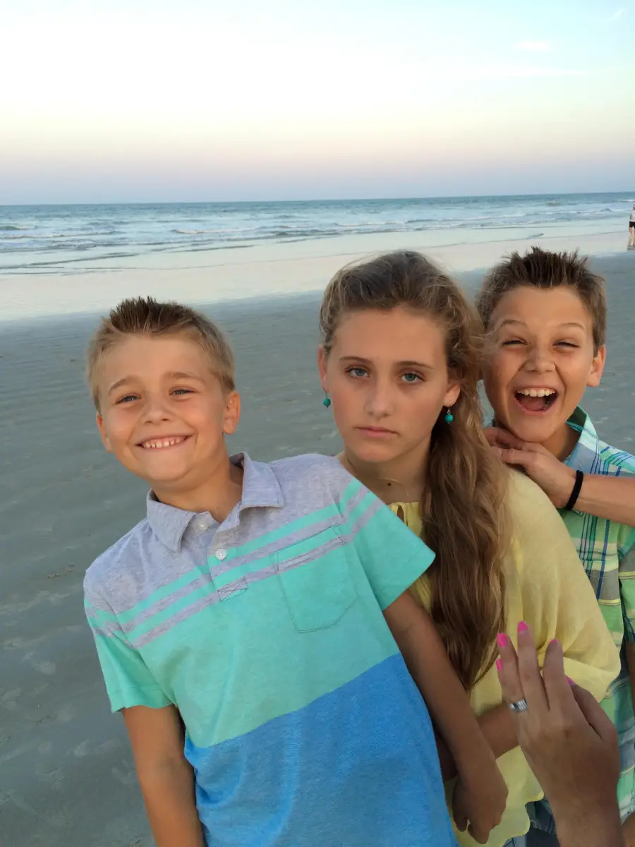 Three children pose on a beach at sunset; two boys smile playfully, while the girl in the middle looks serious. A hand with pink nails appears in the foreground.