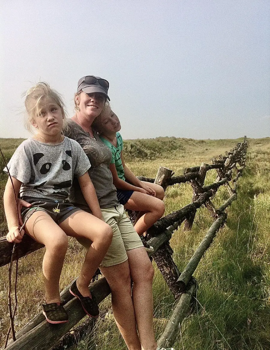 A woman and two children sit on a wooden fence in a grassy field on a sunny day. The children look relaxed, and the woman is smiling.