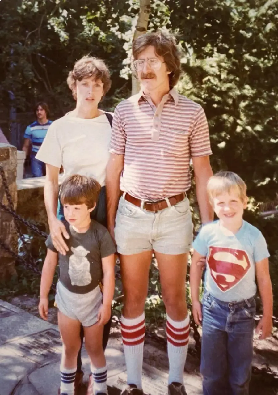 A family poses outdoors: two adults and two boys stand together. One boy wears a Superman shirt, while the man wears striped socks and shorts. Trees and another person are in the background.