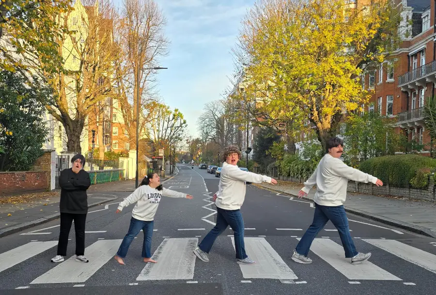 Four people, all dressed similarly, recreate the famous Abbey Road crosswalk pose on a sunny day, with autumn trees and brick buildings lining the street.