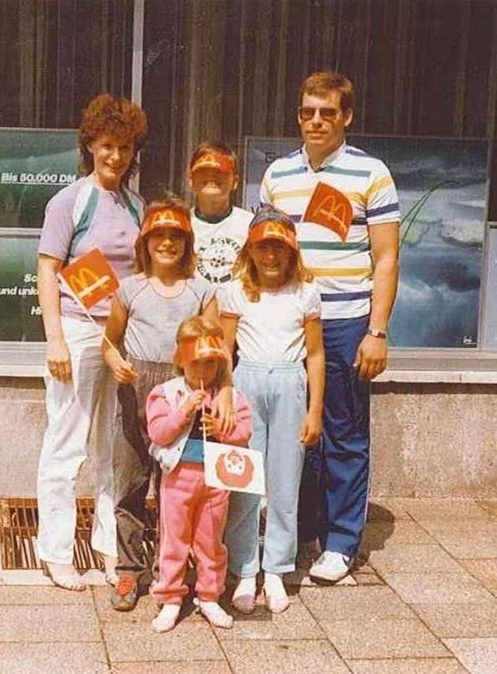 A family of six poses outside. Four children wear McDonald’s hats and hold flags with the logo. The parents stand behind them, smiling. They are dressed in casual 1980s clothing.