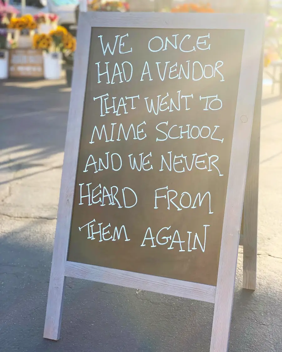A chalkboard sign reads: “We once had a vendor that went to mime school and we never heard from them again.” Flowers are visible in the background.
