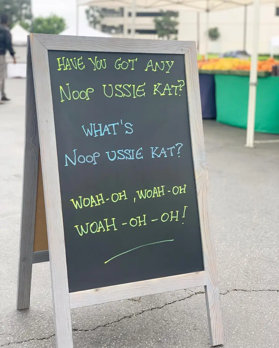 A chalkboard sign at a market with playful text: “Have you got any noop ussie kat? What’s noop ussie kat? Woah-oh, woah-oh, woah-oh-oh!” Fruit stands are visible in the background.