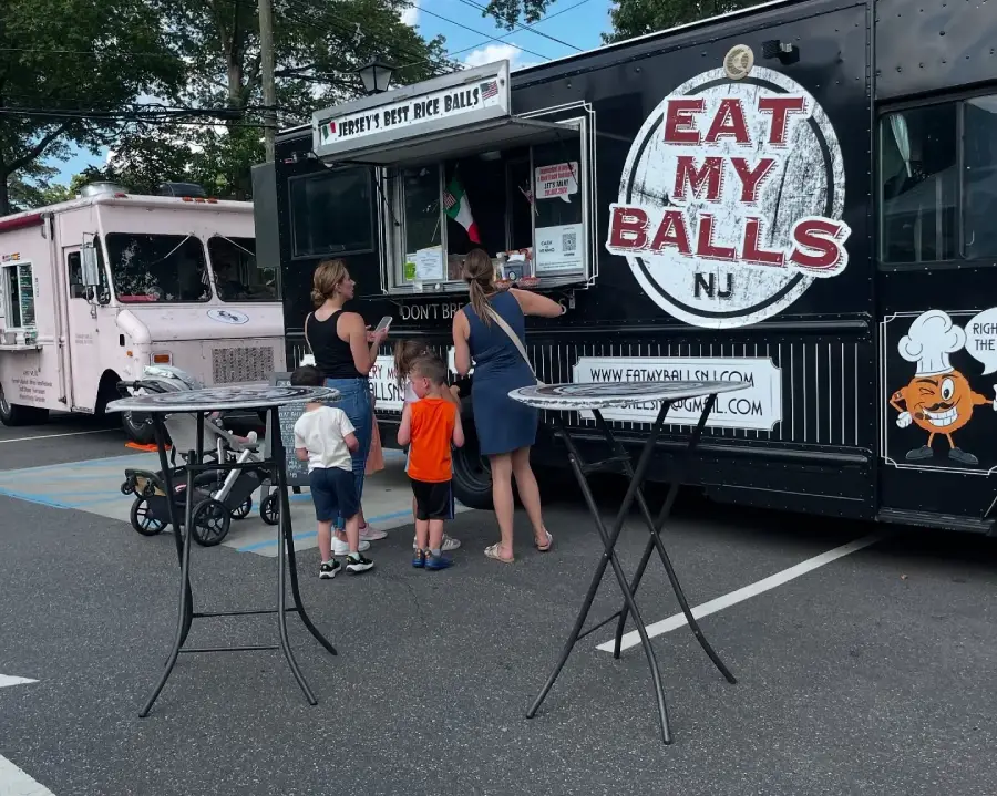 A family orders food at a black food truck labeled “EAT MY BALLS NJ.” Another food truck and two empty tall tables are nearby in a parking lot.