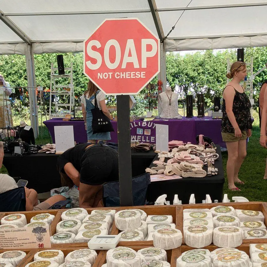 A red sign shaped like a stop sign reads “SOAP NOT CHEESE.” In front, round soaps are displayed. People browse stalls under a white tent at an outdoor market.