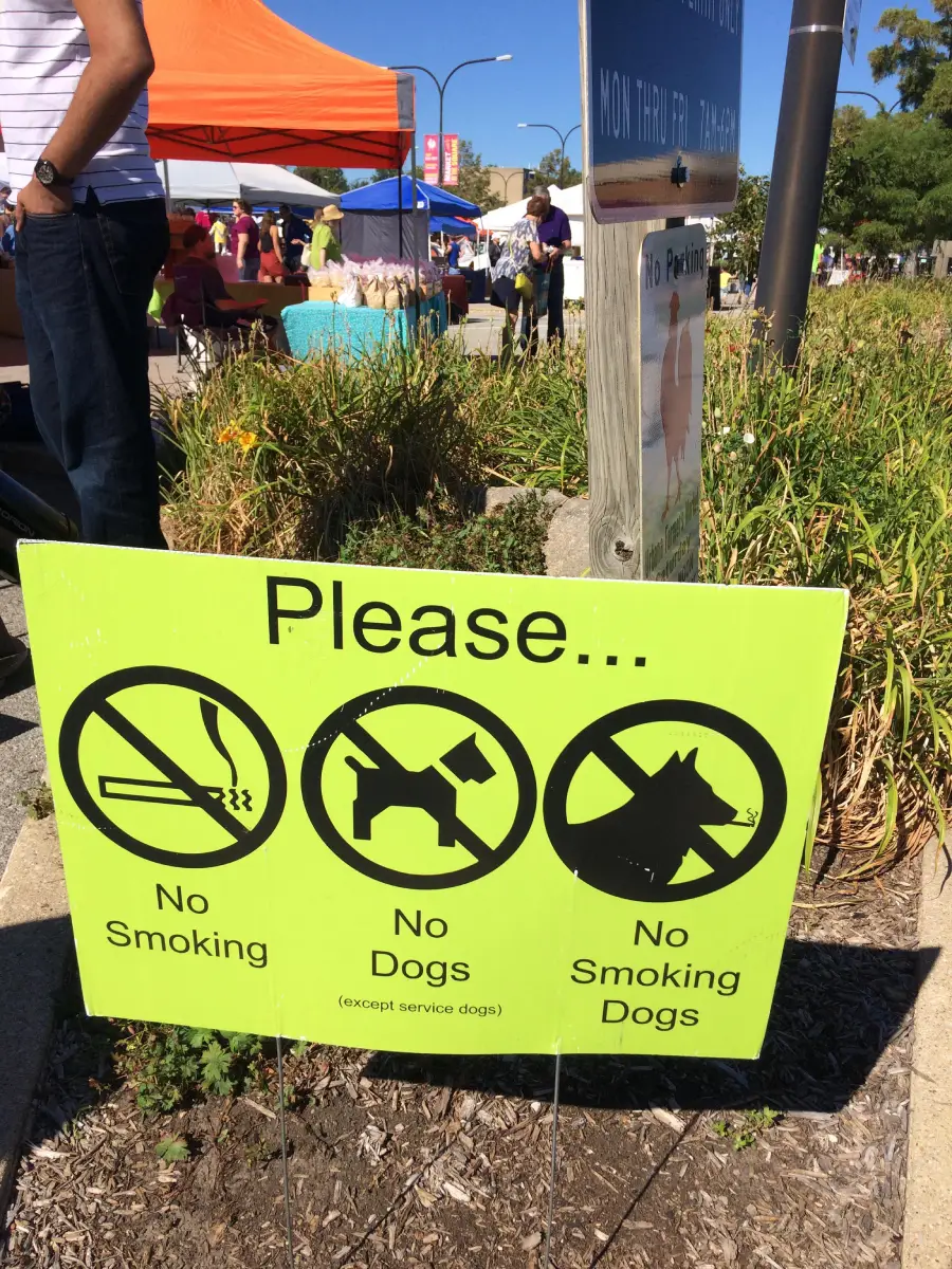 A green sign with icons reads: “Please... No Smoking, No Dogs (except service dogs), No Smoking Dogs,” at an outdoor event with tents and people in the background.