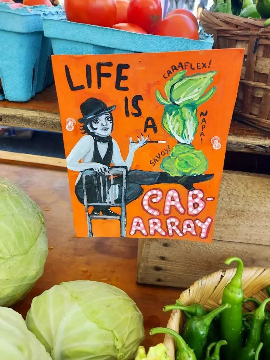 A colorful market sign shows a person in black and white with cabbages. Text reads, “Life is a cab-array,” with playful cabbages labeled “Caraflex,” “Napa,” and “Savoy.” Fresh produce surrounds the sign.