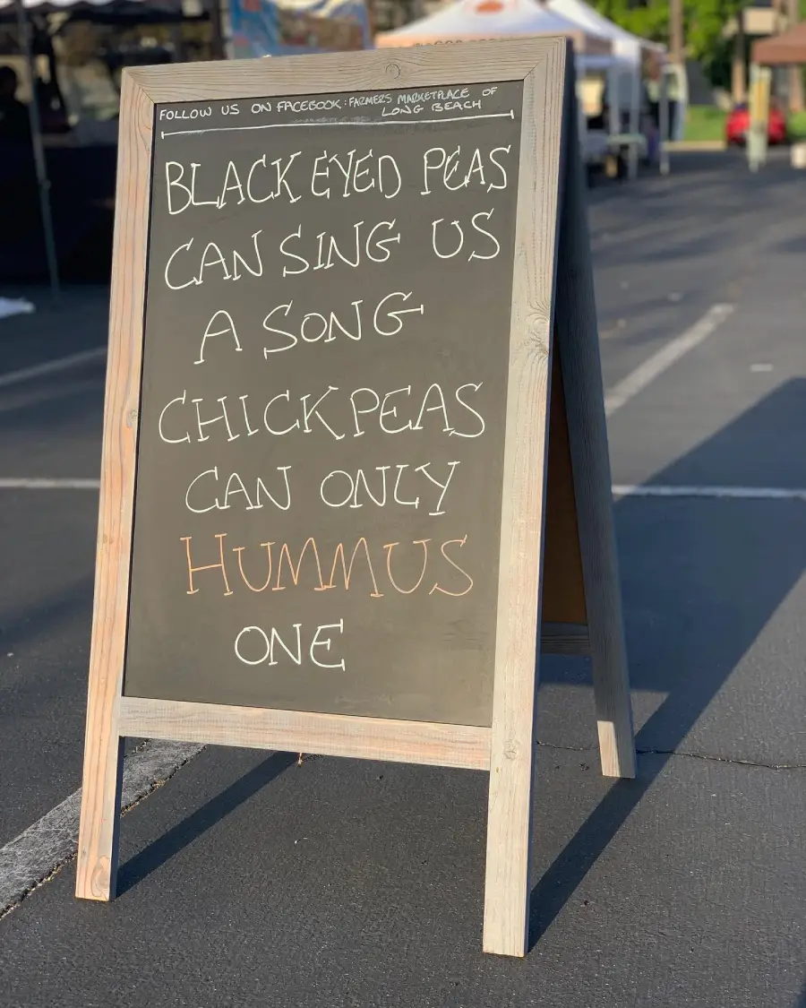 A chalkboard sign reads: “Black Eyed Peas can sing us a song — Chickpeas can only hummus one.” The sign is on a sidewalk at an outdoor market.