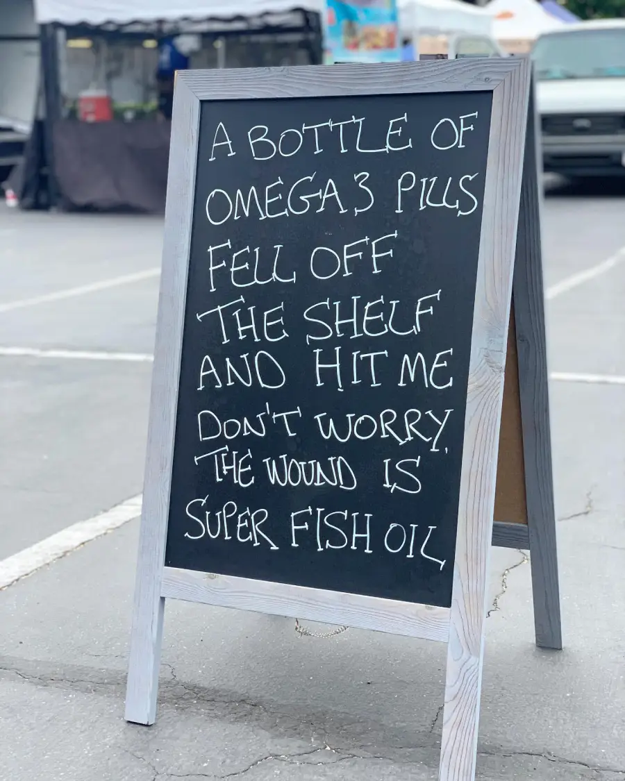 A chalkboard sign with a joke written on it about Omega 3 pills falling and causing a “super fish oil” wound. The sign stands in a parking lot with market stalls and a van in the background.