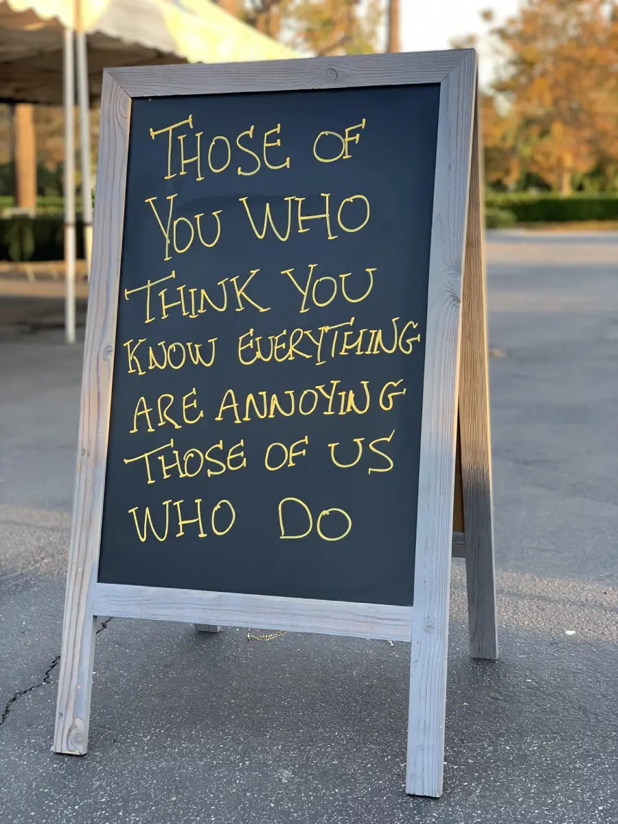 A chalkboard sign reads: “Those of you who think you know everything are annoying those of us who do.” The sign is outdoors on a paved area with some greenery and tents in the background.