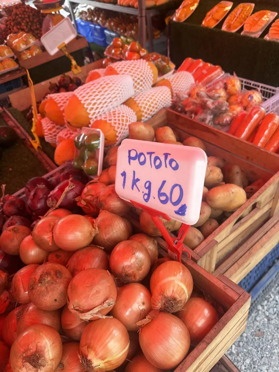 A market stall displays onions and other vegetables. A handwritten sign reading “Pototo 1 kg. 60” is placed among the onions. Other produce is visible in the background.