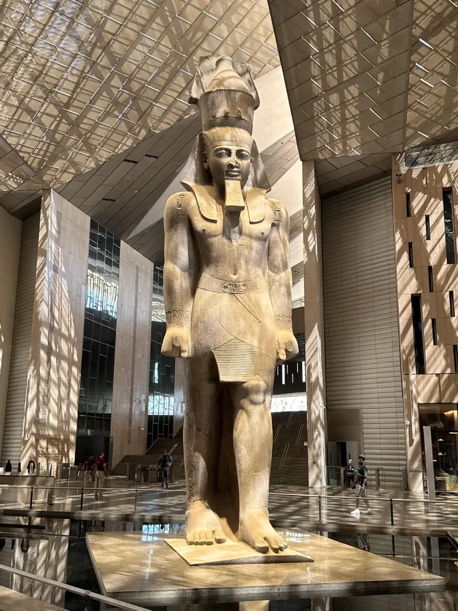 A large ancient Egyptian statue stands in a modern museum atrium, surrounded by glass walls, a patterned ceiling, and reflective floors. People nearby appear much smaller in comparison.