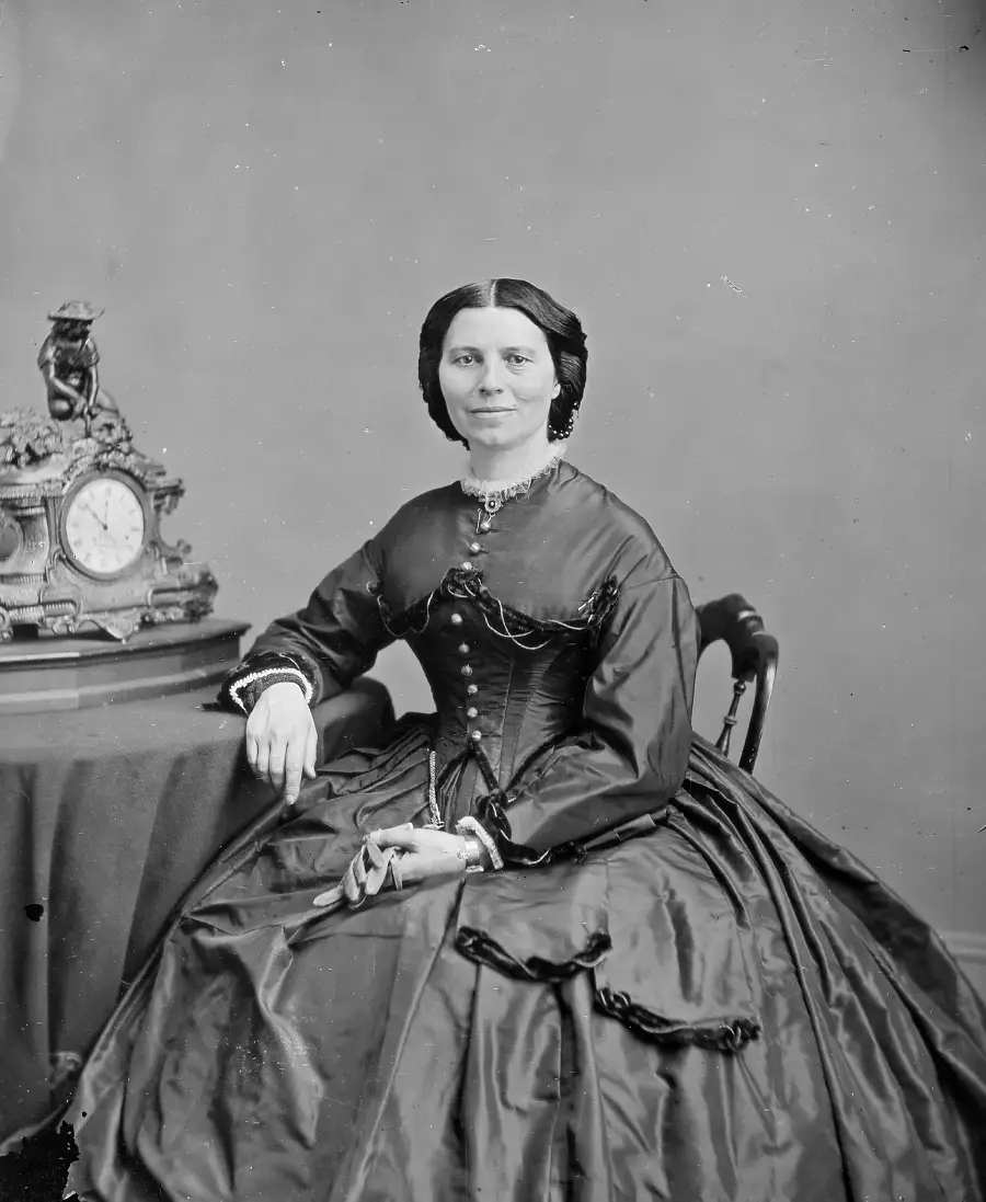 A woman in a dark 19th-century dress sits beside a table with an ornate clock, looking at the camera, her hair parted in the center and styled smoothly.