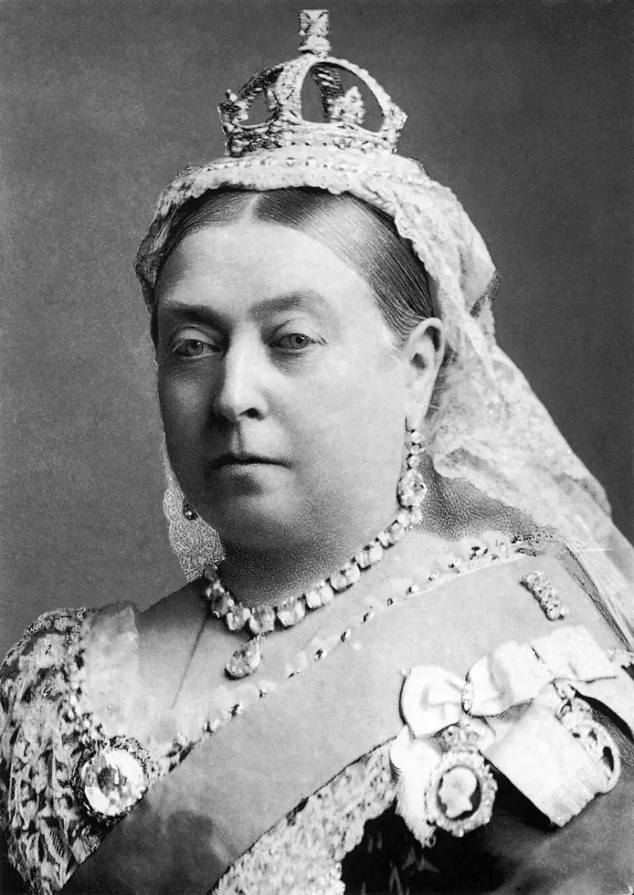 A stern-faced woman in regal attire wearing a crown, ornate jewelry, and a lace veil, posed for a formal black-and-white portrait.