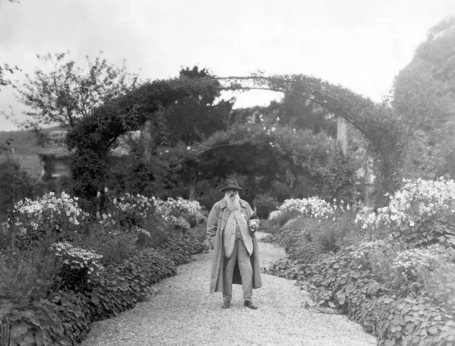 An older man in a long coat and hat stands on a gravel path, surrounded by lush gardens and flowers, beneath an arch covered in greenery.