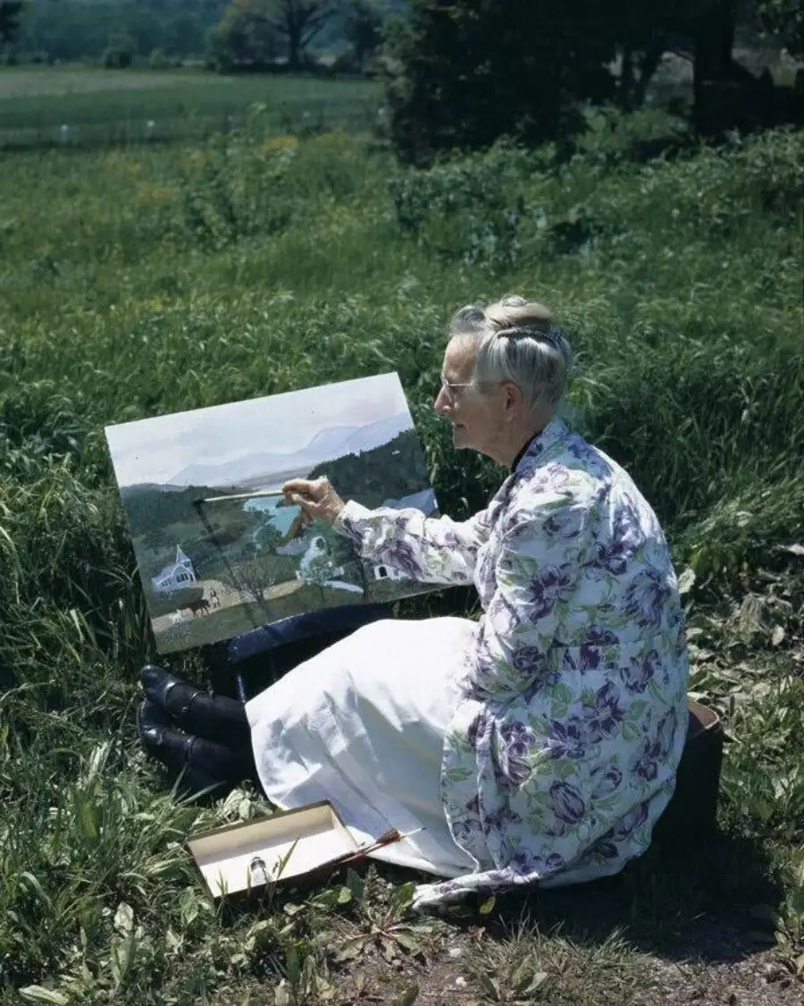 An elderly woman in a floral coat sits on the grass painting a landscape on canvas, capturing trees, hills, and a small white building in the scene before her. Art supplies rest beside her in a box.