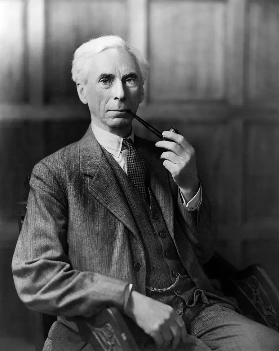 An older man with white hair sits in a chair, wearing a three-piece suit and holding a pipe to his mouth. The background features wooden paneling.