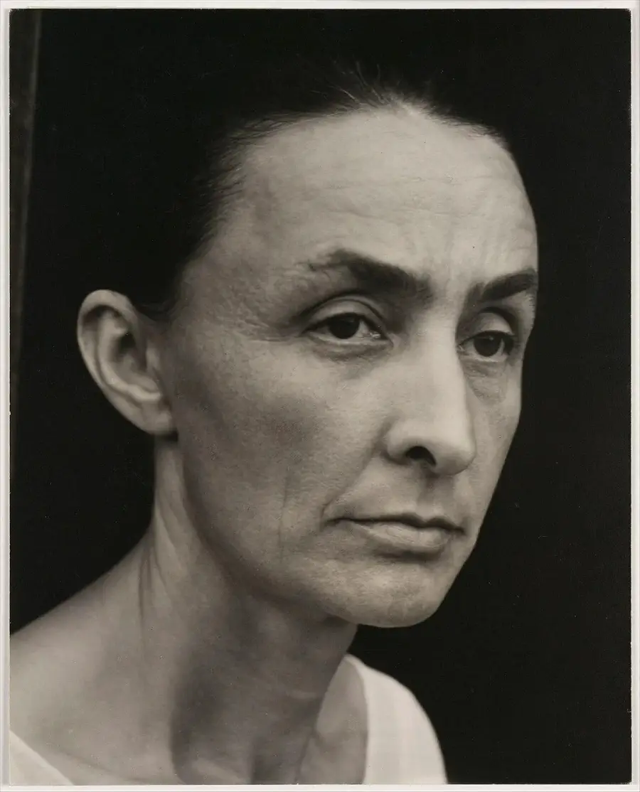 Black and white close-up portrait of a woman with dark hair pulled back, wearing a white top, looking slightly to the side with a serious, contemplative expression.