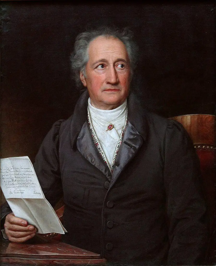 An older man with gray hair sits in a dark coat, holding a handwritten letter and looking to his left. The background is dark, and he is seated in a wooden chair.