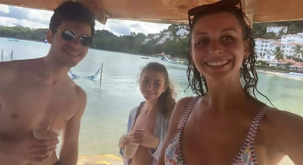 Three young adults in swimsuits smile under a wooden canopy by the water, holding drinks. The background features calm water, distant boats, and buildings on a hillside.