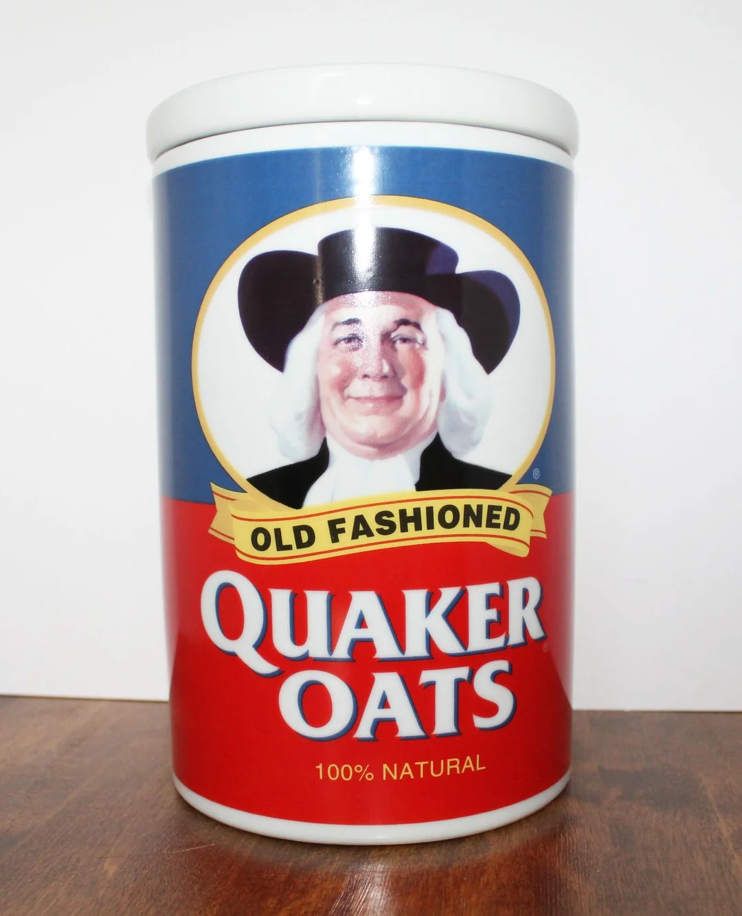 A cylindrical container of Quaker Oats with a smiling person in a black hat and white hair on the label, displaying the words “Old Fashioned Quaker Oats 100% Natural.”