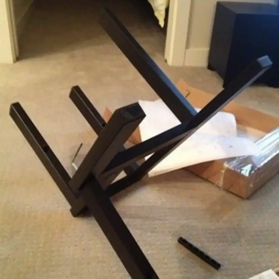 A partially assembled black table lies upside down on a carpeted floor, with instructions and packaging nearby, suggesting a confusing or incorrect assembly attempt.