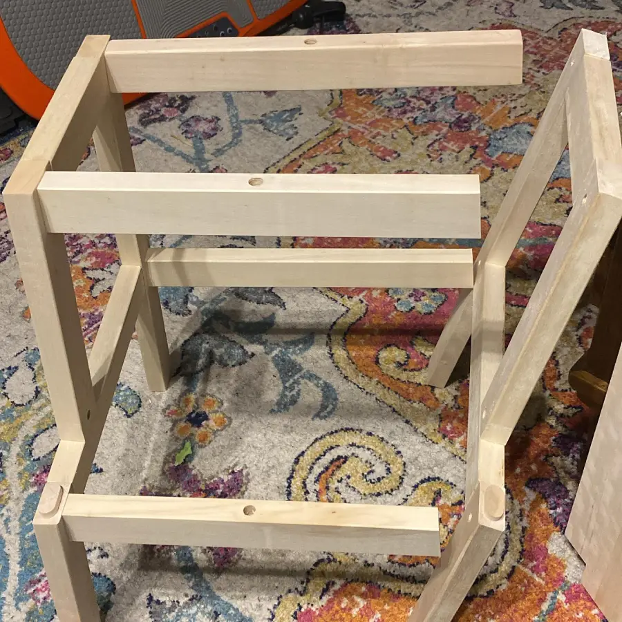 Unfinished wooden frame structure sits on a colorful patterned rug, possibly as part of a DIY furniture or shelving project, with tools and materials visible in the background.