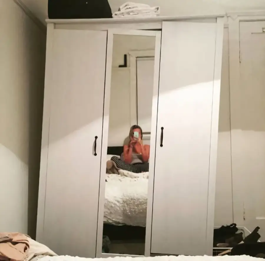 A woman sits on a bed taking a photo in a mirror attached to a white wardrobe. The room is simple with light walls, some clothes visible, and folded towels on top of the wardrobe.