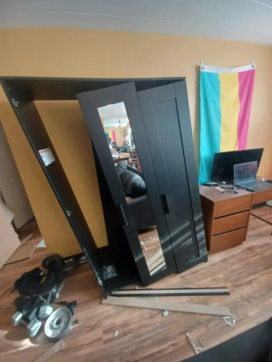 A black wardrobe has collapsed in a room, with doors and panels scattered on the floor. A desk with a computer and a colorful flag hanging on the wall are visible in the background.